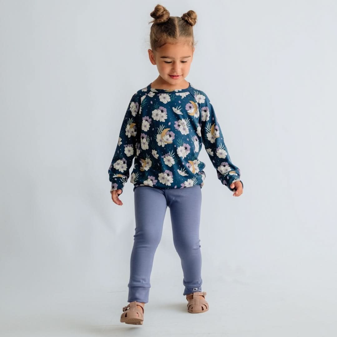 A young child wears the Little & Lively Baby/Kids Bamboo Raglan Pullover | Prints, paired with light purple leggings and light brown shoes, standing against a plain background with hair in two buns, looking slightly downward while walking.