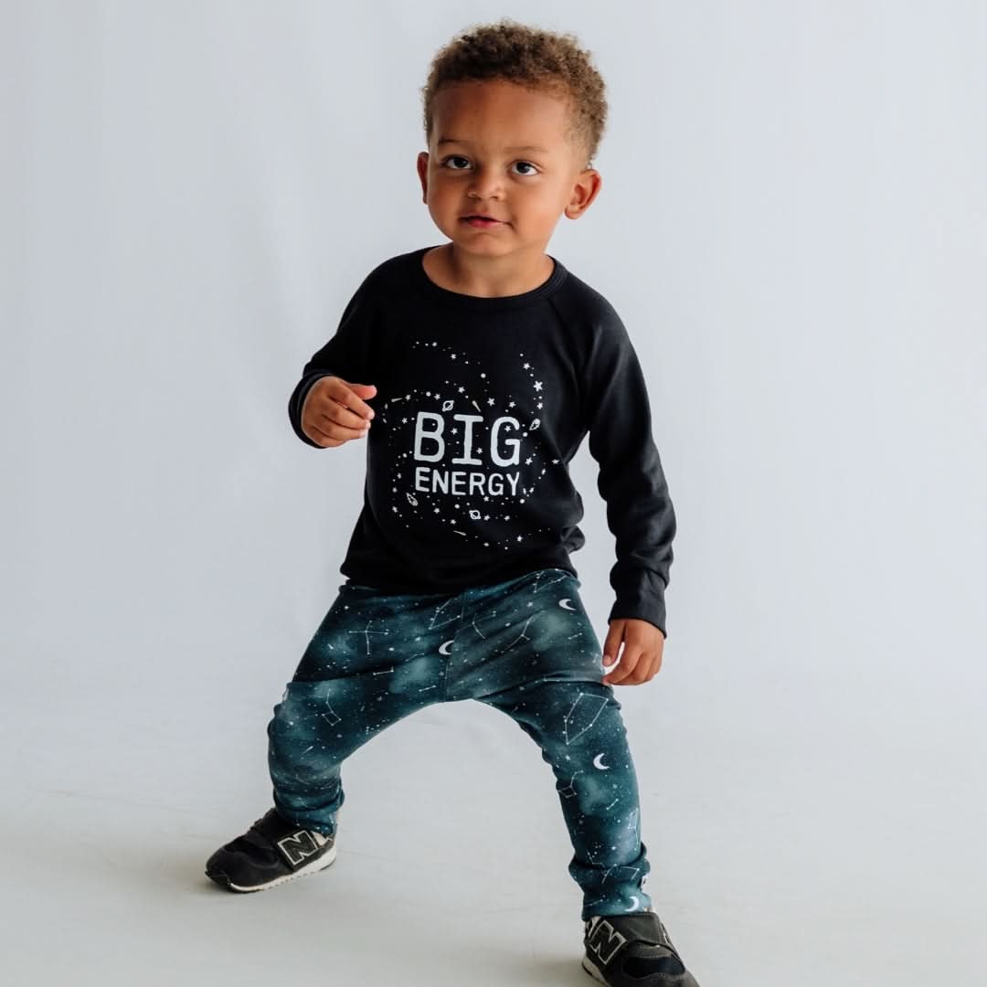 A young child stands against a white background, wearing the Little & Lively Baby/Kids Graphic Bamboo Raglan Sleeve Pullover—perfect playful bedtime wear with "BIG ENERGY" print and paired blue space-patterned pants and black sneakers.