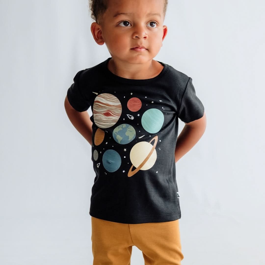 A child stands against a plain backdrop, wearing the Little & Lively Baby/Kids Bamboo Graphic T-shirt with colorful planets and mustard yellow pants, looking slightly upward and to the side.