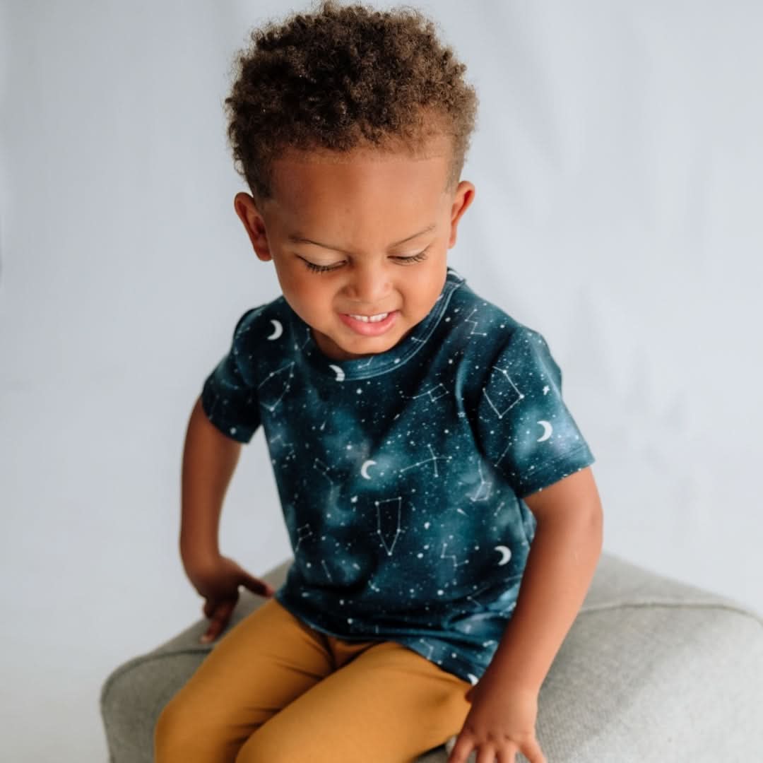 A young child with short curly hair sits on a light gray cushion, wearing the Little & Lively Baby/Kids Bamboo T-shirt with space-inspired prints and mustard yellow pants, looking down and smiling slightly against a light background.