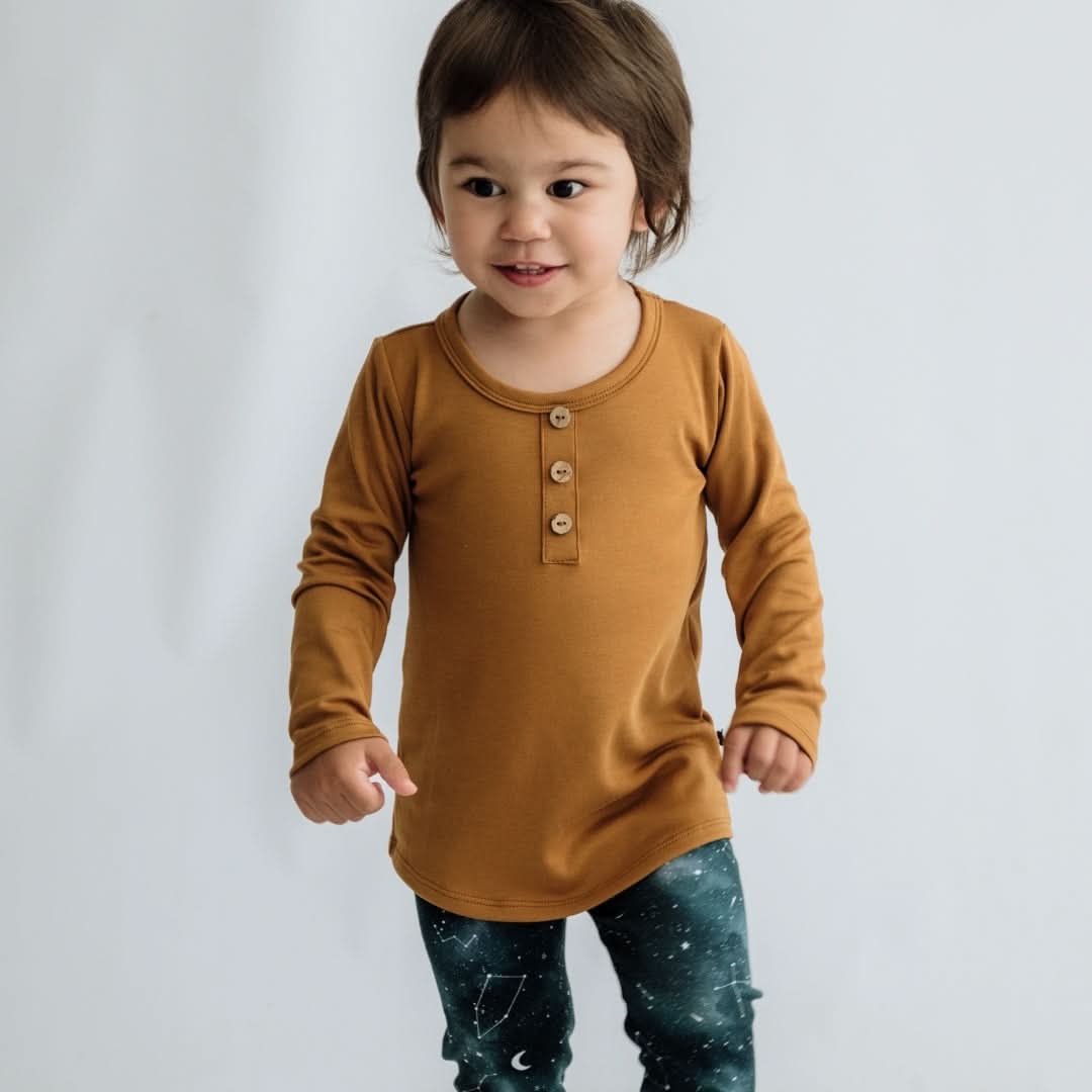 A young child with short brown hair wears the Little & Lively Baby/Kids Bamboo Long Sleeve Henley in mustard and dark leggings with a white star and constellation pattern, standing against a plain light background.