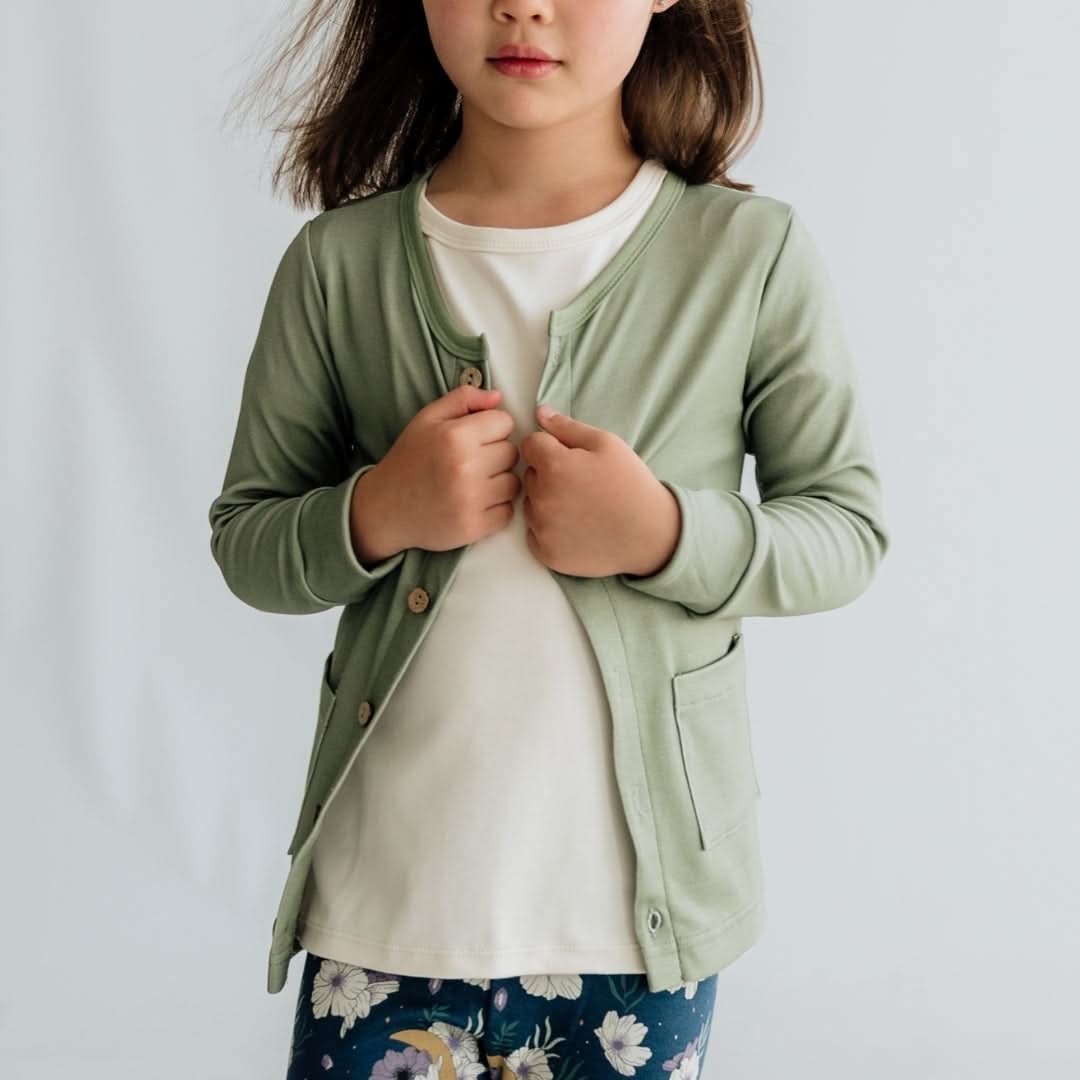 A young girl stands against a plain background, wearing the Kids Bamboo Cardigan by Little & Lively—a light green Soothknit piece with eco-friendly coconut buttons—over a white shirt and floral pants, holding the cardigan closed. Her face is partially out of frame.