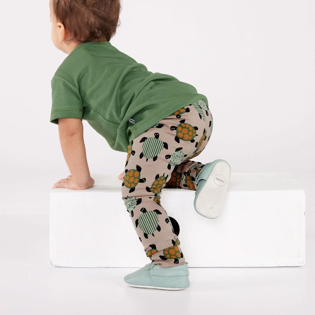 A toddler in a green t-shirt, turtle-print beige pants, and light blue shoes climbs on a white block—an ideal scene to highlight Little & Lively’s Kids Bamboo Leggings for comfy youth outfit pairings.