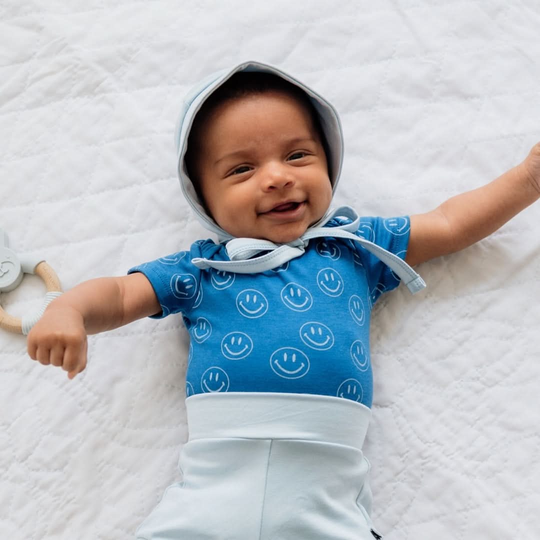 A baby in a blue onesie with smiley faces and the Little & Lively Baby/Kids Bamboo Garden Bonnet lies on a white quilted surface, smiling with arms outstretched. A pacifier clip is visible to the left.