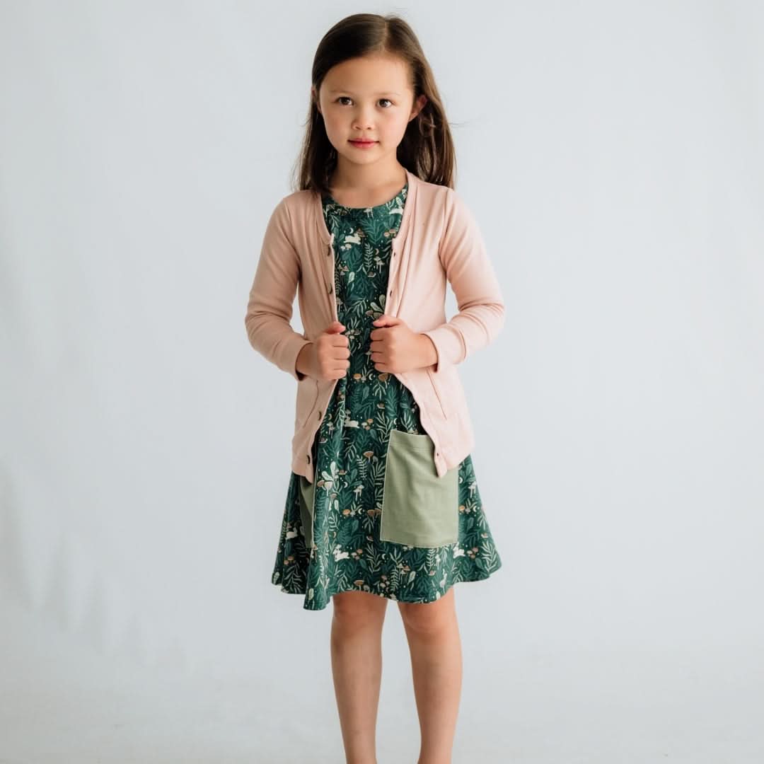 A young girl wears the Little & Lively Baby/Kids Bamboo Clementine Dress with a light green pocket and a light pink cardigan, standing against a plain background while holding the edges of her cardigan and gazing slightly to the side.