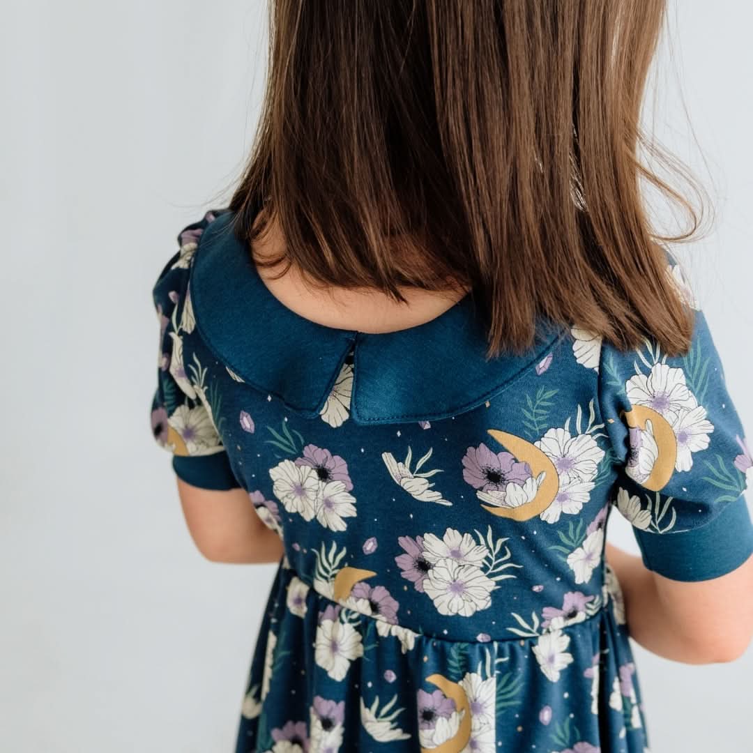 A young girl with straight brown hair is seen from behind, wearing the Little & Lively Kids Bamboo Penelope Dress—a blue floral day dress with puff sleeves and a Peter Pan collar—set against a plain, light-colored background.