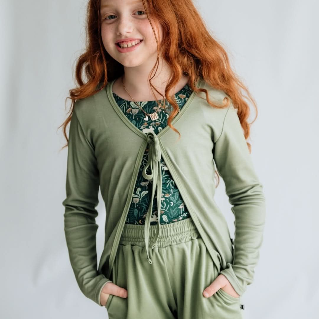A young girl with long red hair smiles with her hands in the pockets of her light green pants, wearing the Little & Lively Youth Celeste Bamboo Cardigan over a dark green patterned shirt. The background is plain and light-colored.