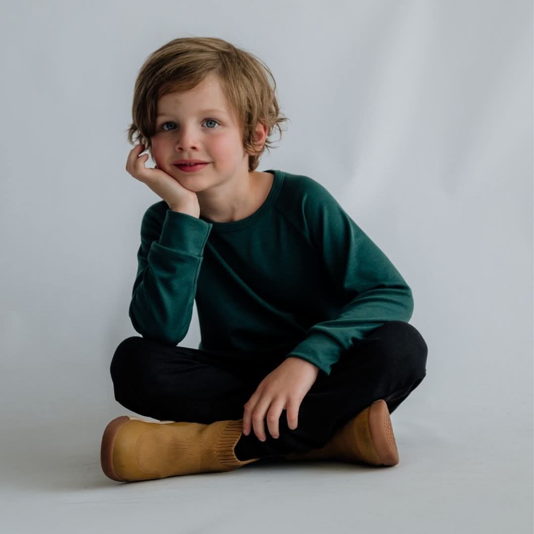 A young child with light brown hair sits cross-legged on the floor against a plain light background, wearing the Little & Lively Youth Bamboo Raglan Pullover in dark green, paired with black pants and tan boots, and resting their face on one hand.