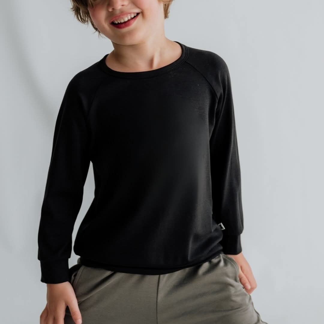 A light brown-haired child wears the Little & Lively Youth Bamboo Raglan Pullover—black, long-sleeved and breathable—paired with gray pants, posing against a light gray background with a slight smile and one hand in their pocket.