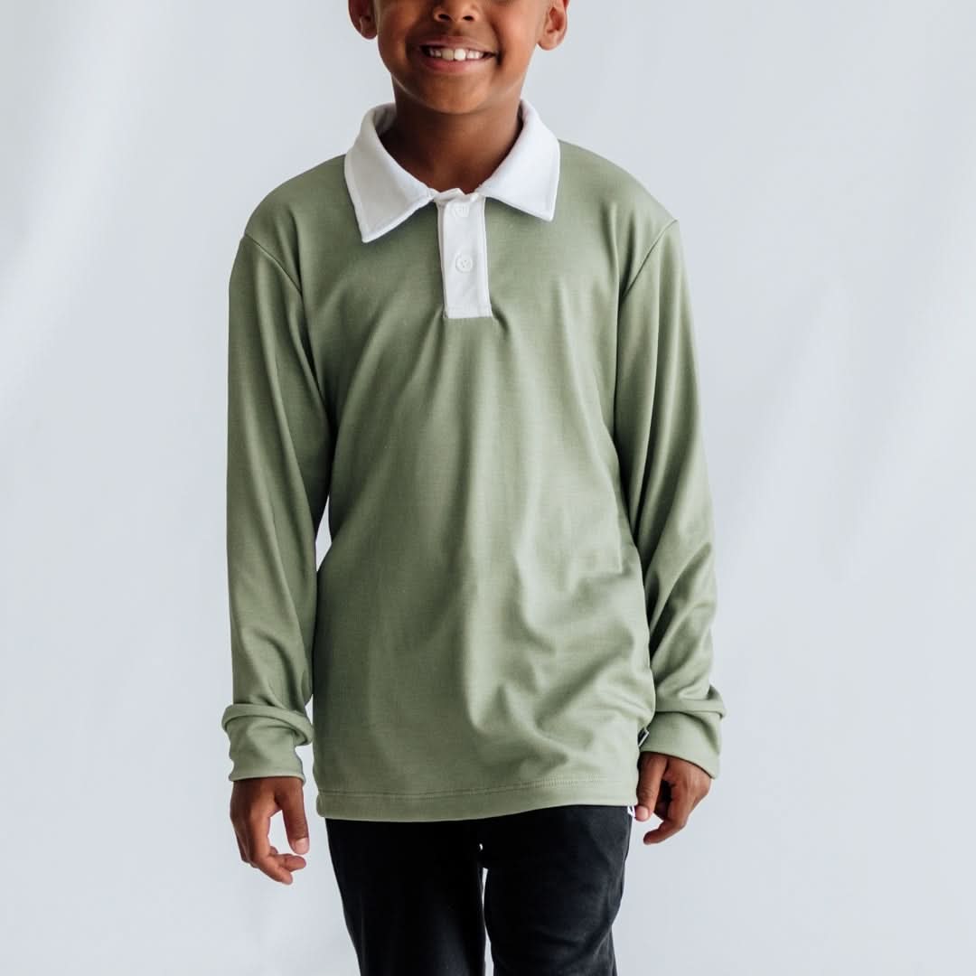 A child stands smiling in a Little & Lively Youth Bamboo Style Rugby Shirt with a white collar and black pants, against a plain light background.