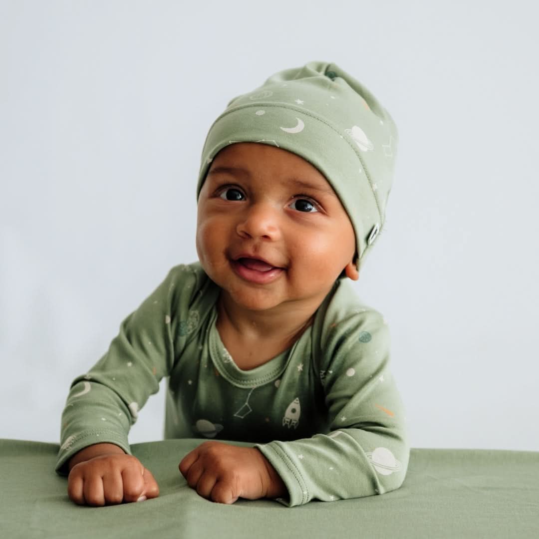 A baby in a Little & Lively Baby/Kids' Bamboo Beanie and a matching green space-themed outfit sits at a green table, smiling and looking to the side.