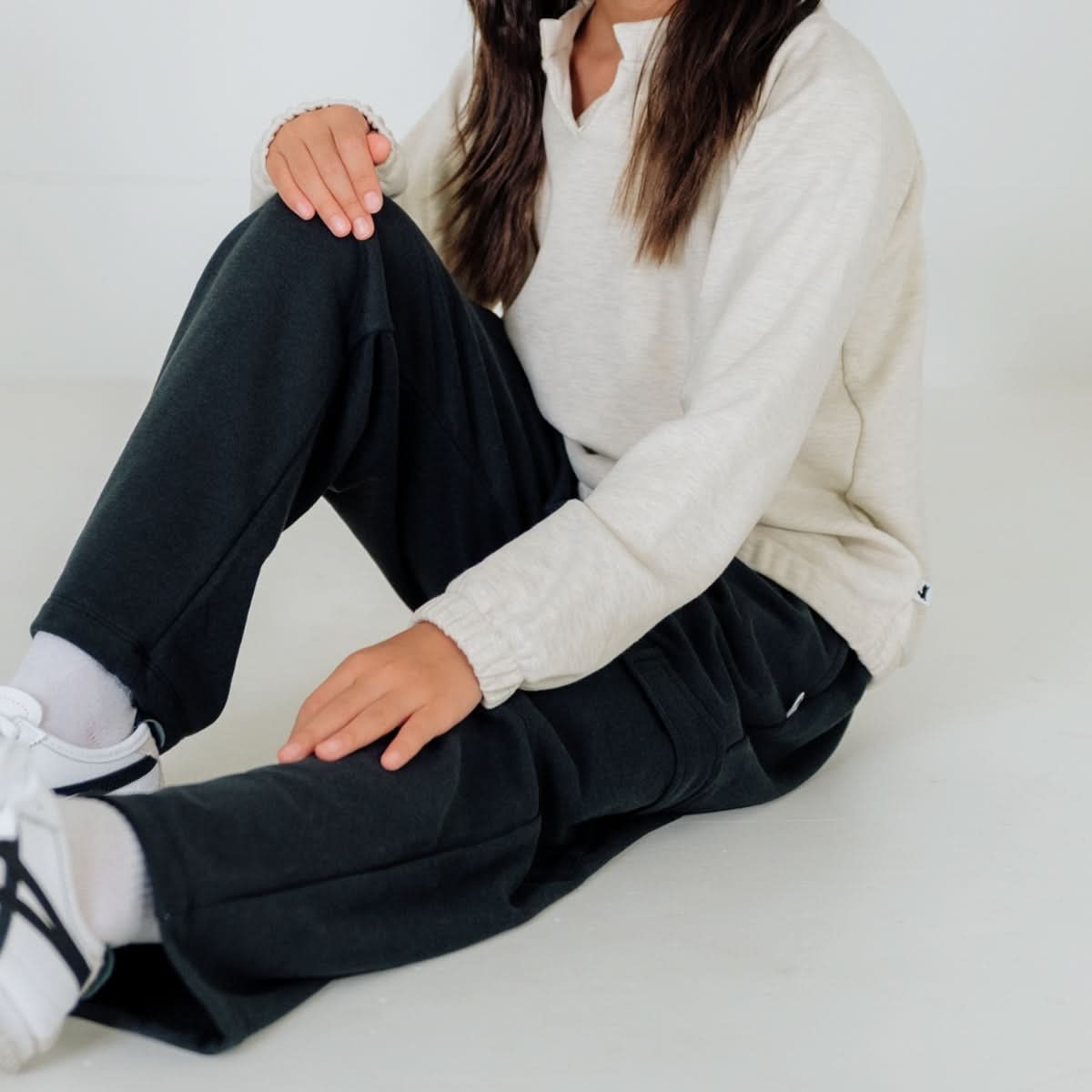A person with long brown hair sits on the floor wearing a beige long-sleeve shirt, Little & Lively Youth Bamboo Fleece Cargo Pants, white socks, and white sneakers. The individual’s face is not visible.