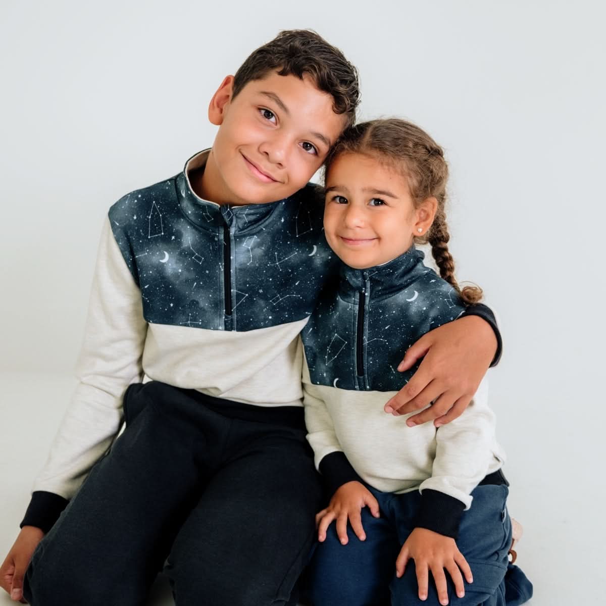 Two children sit close together in matching Little & Lively Kids Bamboo Fleece Half-Zip tops with dark, star patterns. The older child hugs the younger, both looking at the camera against a plain background. Ethically made in Canada.