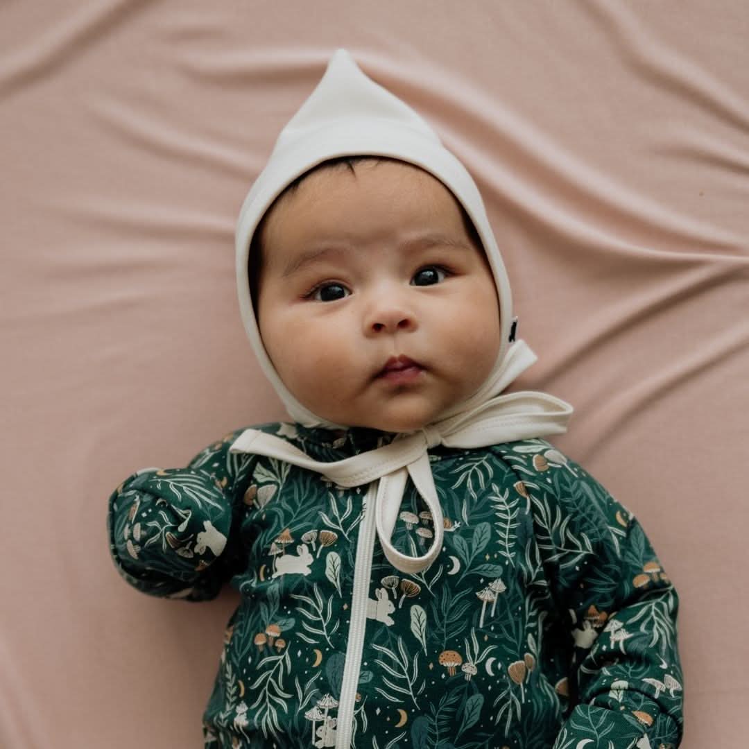 A baby wearing the Little & Lively Bamboo Baby Pixie Bonnet and a green nature-themed outfit lies on a light pink blanket, gazing upward with a neutral expression.