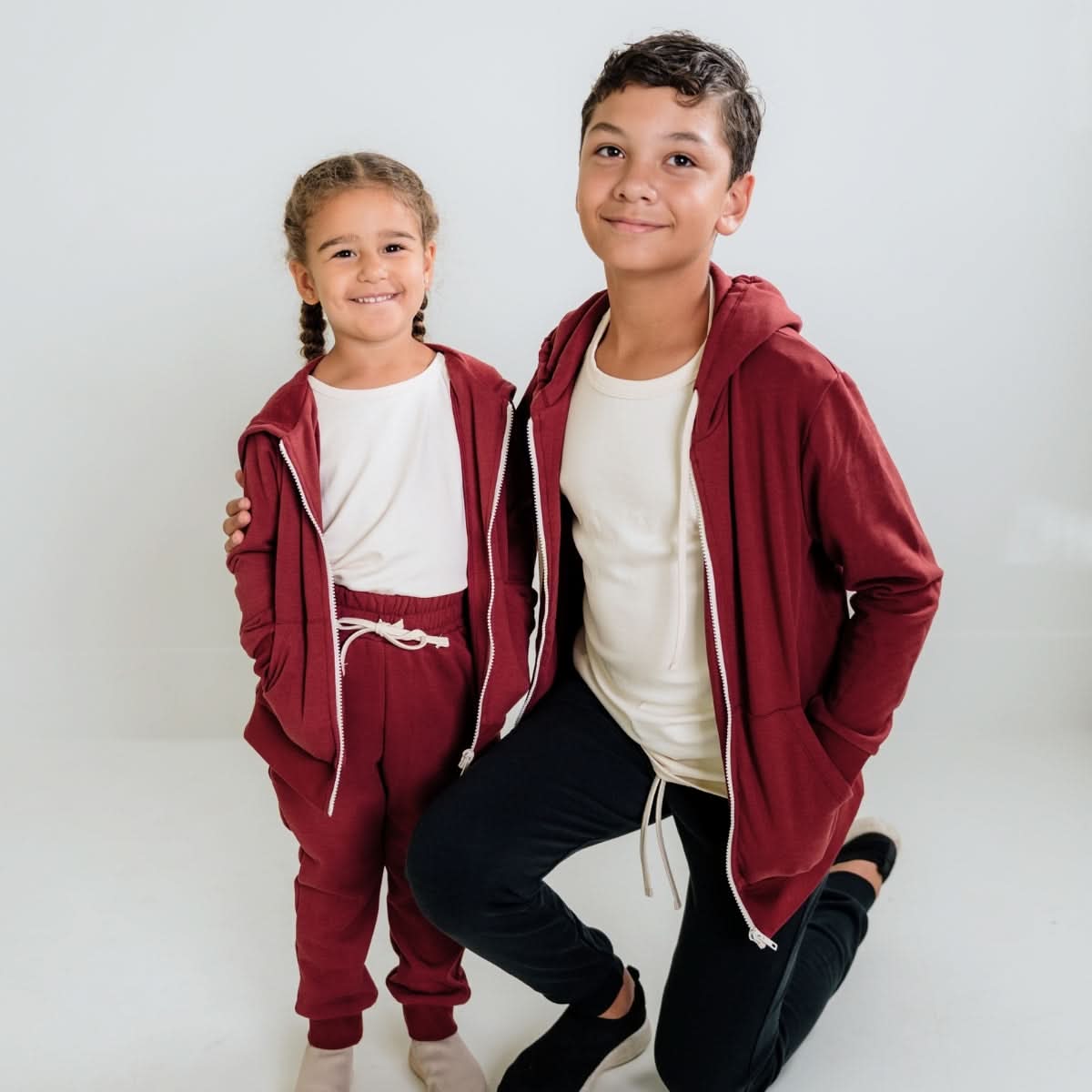 Two kids wear matching Little & Lively Kids Bamboo Fleece Zip-Up Hoodies, made with cozy SootheKnit fabric. One child in maroon pants with braids stands beside an older child in black pants. Ethically made in Canada.