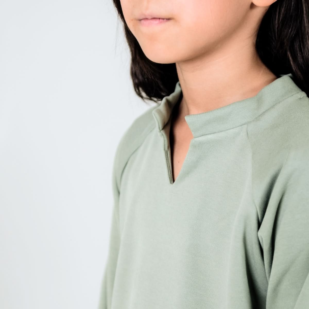 A child with long dark hair wears the Little & Lively Youth Bamboo Fleece Collared Pullover in sage green, ethically made in Canada. Shown from shoulders up against a light background, only their lower face and upper torso are visible.
