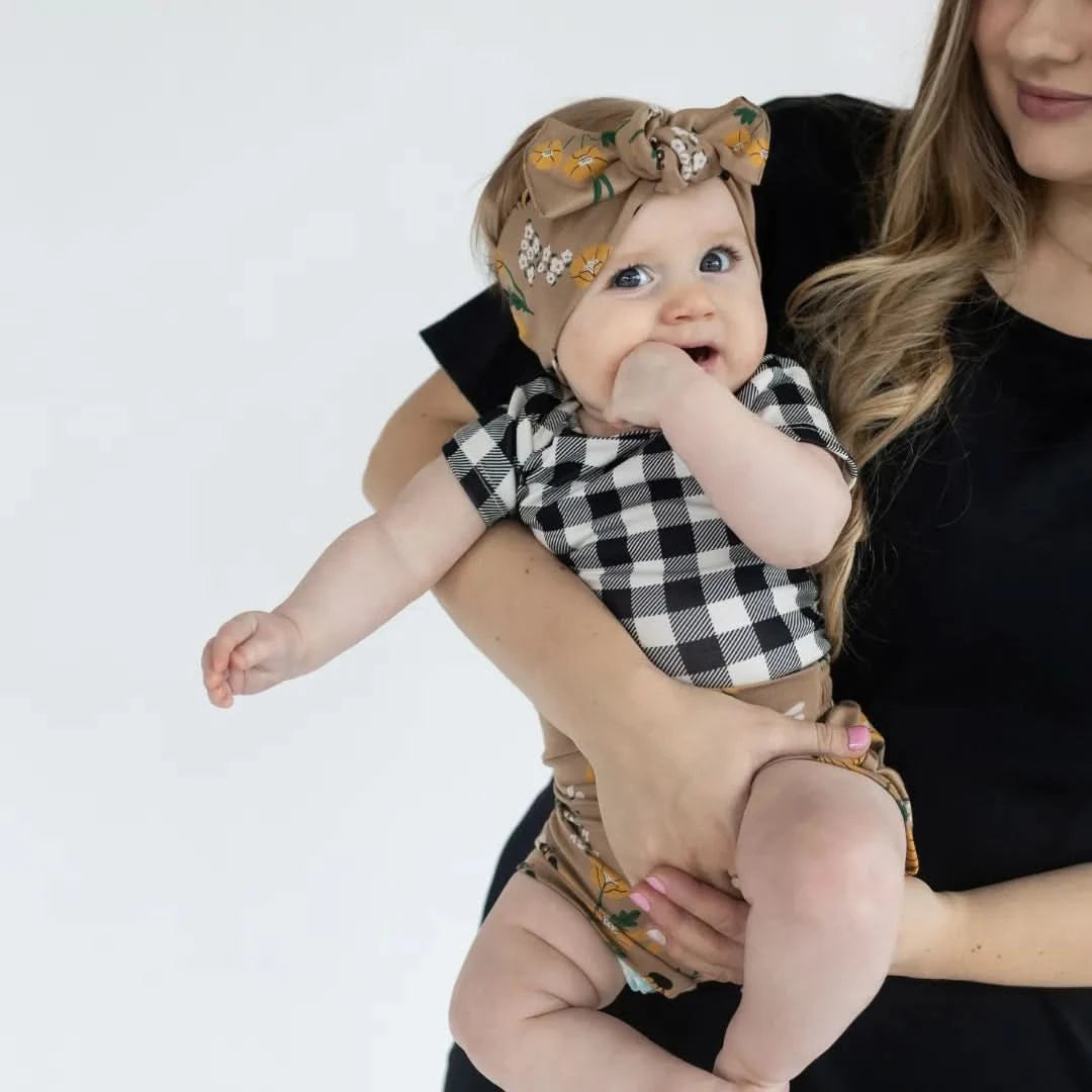 A woman in a black shirt holds a baby wearing Little & Lively’s Baby Bamboo High-Waisted Shorties in black and white checkered print and a brown floral headband, making an adorable addition to any baby wardrobe.