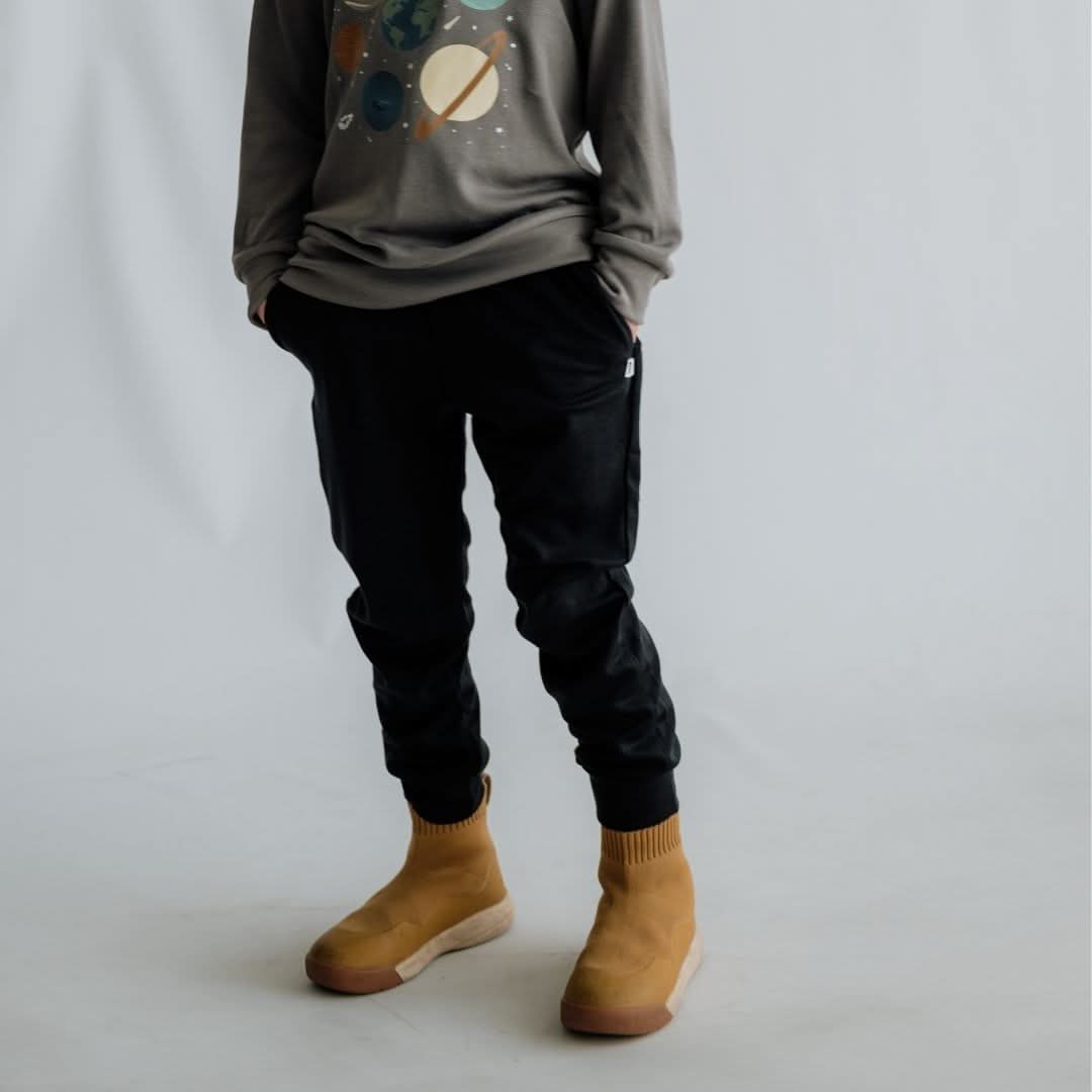 A person stands against a plain light background, wearing black Little & Lively Youth Bamboo Lightweight Joggers, a long-sleeve shirt with a planets design, and tan slip-on boots. The head and upper body are not visible.
