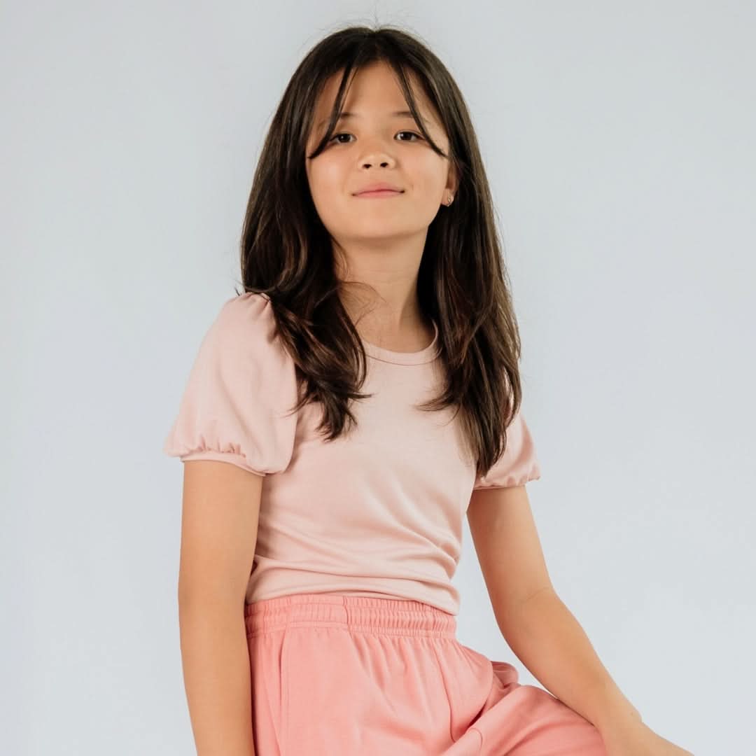 A young girl with long dark hair wears the Little & Lively Youth Bamboo Puffed Sleeve T-shirt in pink and matching pants, seated against a neutral backdrop.