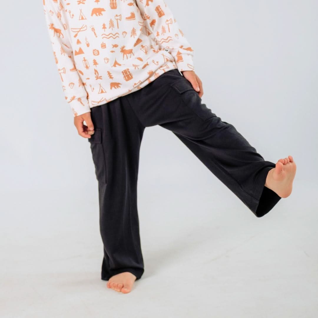 A child in a long-sleeve shirt with brown patterns and Little & Lively Kids Bamboo Cargo Pants with an adjustable waist stands barefoot on one leg, the other slightly lifted, against a plain light background.