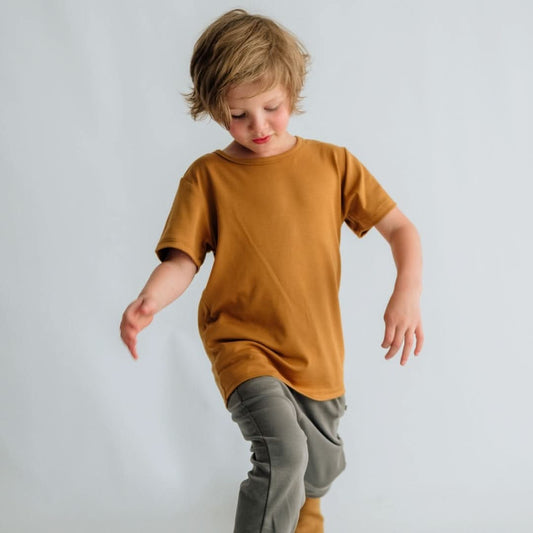 A young child with light brown hair wears the Little & Lively Youth Bamboo T-shirt in mustard and gray pants, standing against a plain light background while looking down and lifting one leg.