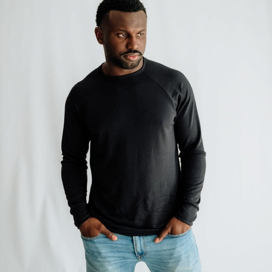 A man stands with his hands in his pockets against a plain white background, wearing light blue jeans and a black Little & Lively Adult Unisex Bamboo Raglan Pullover.
