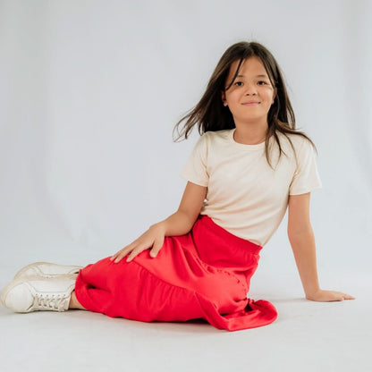A young girl with long brown hair sits on the floor against a white background, wearing a cream t-shirt, white sneakers, and the Little & Lively Youth Bamboo Aria Skirt in bright red.