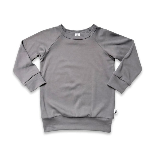 The Little & Lively Youth Bamboo Raglan Pullover is a plain, long-sleeve light gray sweatshirt with ribbed cuffs and waistband, shown flat on a white background. A small white tag with a black logo accents the bottom hem—perfect breathable kids clothing.